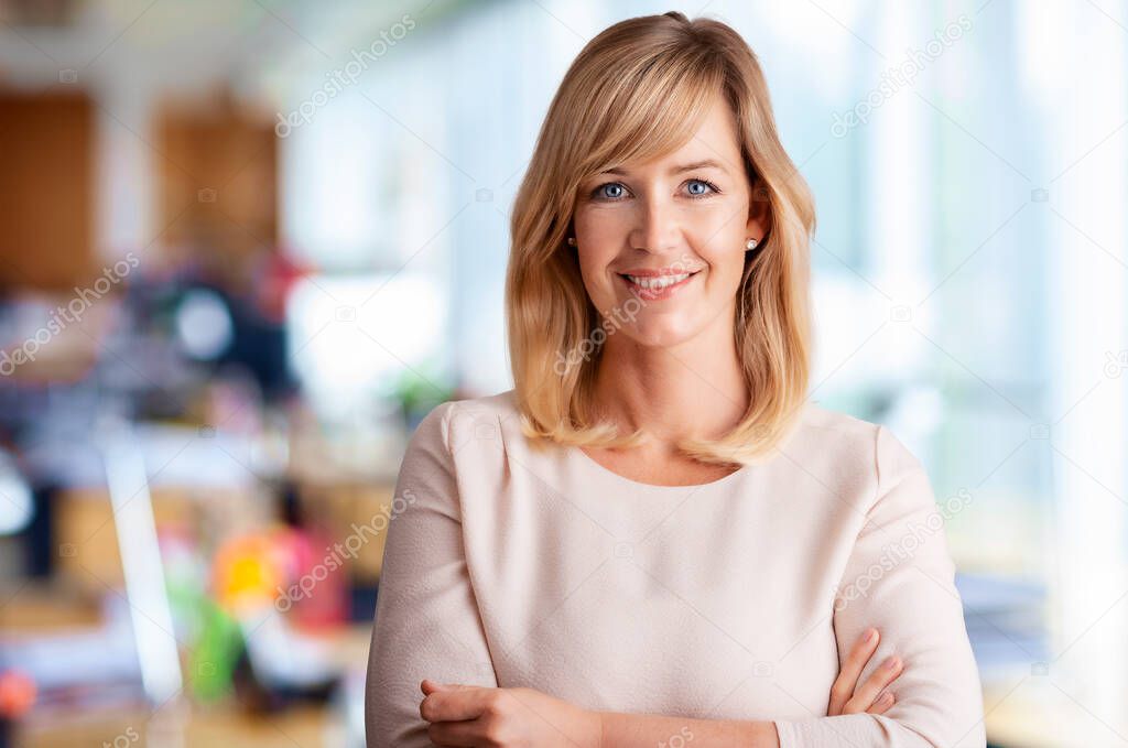 Portrait of smiling blond haired businesswoman while standing with arms crossed at the office.