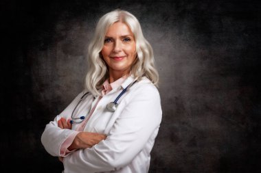 Tecrit edilmiş koyu arkaplan kollarıyla katlanmış yaşlı bayan doktorun stüdyo portresi.. 