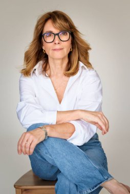 Sarışın kadın izole edilmiş arka planda güvenle oturuyor ve kameraya bakıyor. Beyaz gömlek ve kot pantolon giyen orta yaşlı bir kadın. Boşluğu kopyala. 