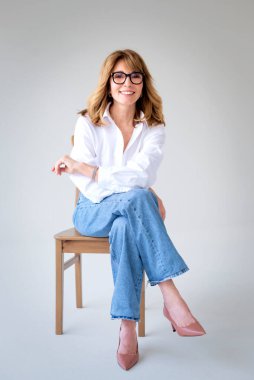 Sarışın kadın izole edilmiş arka planda güvenle oturuyor ve kameraya bakıyor. Beyaz gömlek ve kot pantolon giyen orta yaşlı bir kadın. Uzayı kopyala Tam uzunluk. 