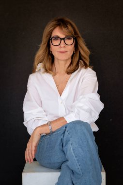 Sarışın kadın, izole edilmiş siyah arka planda güvenle oturuyor ve kameraya bakıyor. Beyaz gömlek ve kot pantolon giyen orta yaşlı bir kadın. Boşluğu kopyala. 