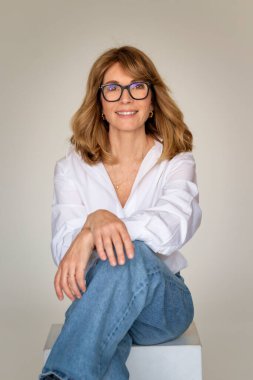 Sarışın kadın izole edilmiş arka planda güvenle oturuyor ve kameraya bakıyor. Beyaz gömlek ve kot pantolon giyen orta yaşlı bir kadın. Boşluğu kopyala. 