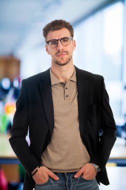 Modern bir ofiste tek başına dururken gülümseyen kendinden emin genç bir iş adamının portresi. Ceket ve gömlek giyen genç bir girişimci. Yakın çekim..