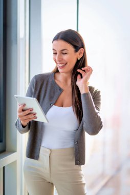 Kendine güveni olan profesyonel bir kadın geniş pencereli modern bir ofiste duruyor. Esmer, iş kıyafeti giyen ve elinde bir dokunmatik ped tutan bir iş kadını. Şirket hayatı. 