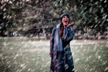Kar yağarken dışarıda parkta duran mutlu kadın. Esmer kadın, şapka ve atkı takıyor ve gülümsüyor.. 