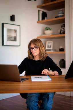 Kendine güveni tam orta yaşlı bir kadın modern bir ofis masasında rahatça oturuyor. Siyah kazak giyen ve iş için laptop kullanan çekici bir kadın.. 