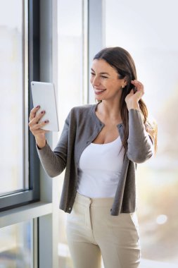 Kendine güveni olan profesyonel bir kadın geniş pencereli modern bir ofiste duruyor. Esmer, iş kıyafeti giyen ve elinde bir dokunmatik ped tutan bir iş kadını. Şirket hayatı. 