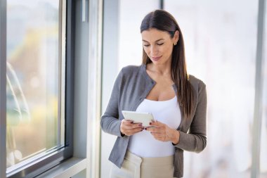 Kendine güveni olan profesyonel bir kadın geniş pencereli modern bir ofiste duruyor. Esmer, iş kıyafeti giyen ve elinde bir dokunmatik ped tutan bir iş kadını. Şirket hayatı. 