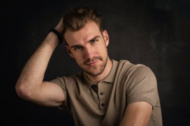 Tecrit edilmiş arka planda oturan yakışıklı bir gencin yakın çekimi. Uzayı kopyala Kendine güvenen erkek polo gömlek giyiyor ve düşünceli görünüyor. Eller başa.. 