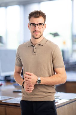 Modern bir ofiste dururken gülümseyen kendinden emin genç bir işadamının portresi. Genç girişimci iş kıyafeti giyer. Yakın çekim..