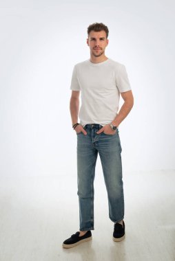 Sıradan kıyafetler giyen ve izole edilmiş beyaz arka planda duran sakallı bir gencin portresi. Beyaz tişört ve kot pantolon. Stüdyo çekimi, kopyalama alanı. Tam uzunluk.