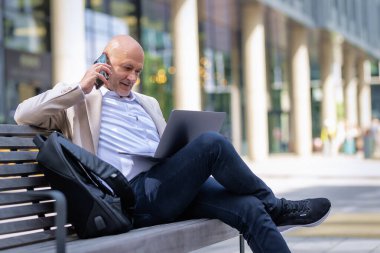 Sokakta bir bankta oturan orta yaşlı bir adam telefon kullanıyor. Gündelik kıyafetler giyen kendinden emin bir erkek..