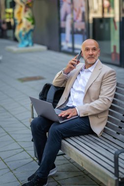Sokakta bir bankta oturan orta yaşlı bir adam telefon kullanıyor. Gündelik kıyafetler giyen kendinden emin bir erkek..
