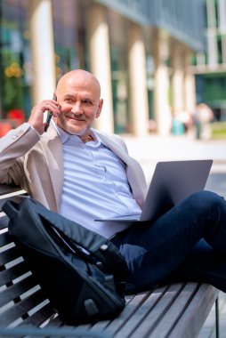 Sokakta bir bankta oturan orta yaşlı bir adam telefon kullanıyor. Gündelik kıyafetler giyen kendinden emin bir erkek..