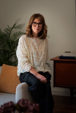 Güzel, orta yaşlı bir kadının rahat evinde koltukta oturduğu uzun bir çekim. Sarı saçlı kadın gömlek ve pantolon giyiyor..