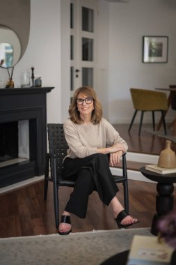 Güzel, orta yaşlı bir kadının rahat evinde koltukta oturduğu uzun bir çekim. Sarı saçlı kadın gömlek ve pantolon giyiyor..