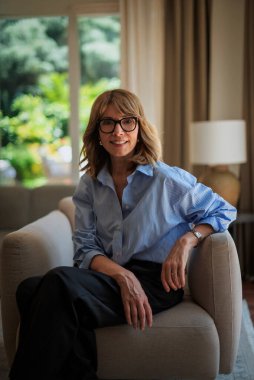 Modern evinde rahat bir koltukta oturan orta yaşlı güzel bir kadının fotoğrafı. Kendinden emin kadın gömleği ve pantolonu var ve kameraya gülümsüyor..
