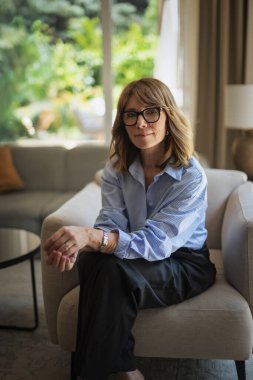 Modern evinde rahat bir koltukta oturan orta yaşlı güzel bir kadının fotoğrafı. Kendinden emin kadın gömleği ve pantolonu var ve kameraya gülümsüyor..