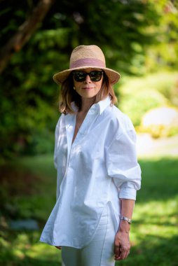 Arka bahçede duran orta yaşlı, sarışın bir kadının portresi. Güneş şapkası ve beyaz gömlek giyen ve dışarıda dinlenen kendine güvenen bir kadın..