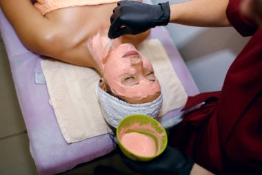 Esmer bir kız spa salonunda prosedürlere tabi tutuluyor, siyah eldivenli bir güzellik uzmanı pembe bir maskeyi kalın bir tabaka içinde uyguluyor, derisini pislikten temizliyor, yüzünü izliyor.