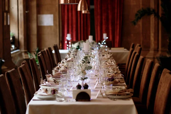A long set table in the restaurant with many transparent glasses, napkins and tableware decorations. The dark interior of the restaurant in the style of a medieval fortress is ready to welcome guests