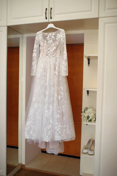 白いウェディングドレスは ウェディングアクセサリー 花嫁の花束の隣のハンガーに掛けられています 花嫁は彼女の服を着る用意ができている 完璧な結婚式のためのウェディングアクセサリーのコンセプト — ストック写真