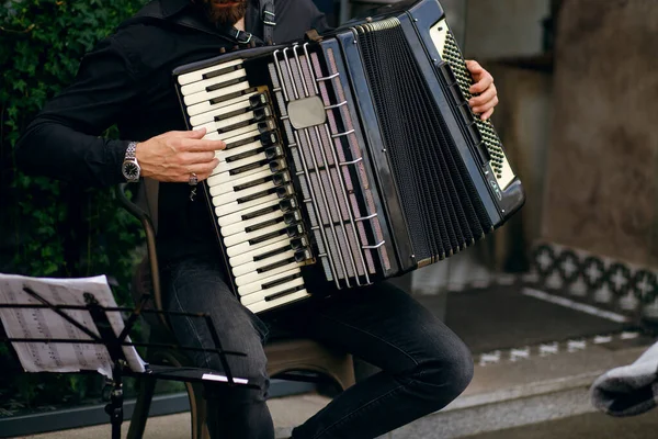 Street musician playing accordion, stylish man cheers up with beautiful music and earns money traveling the world, young man plays near an expensive restaurant