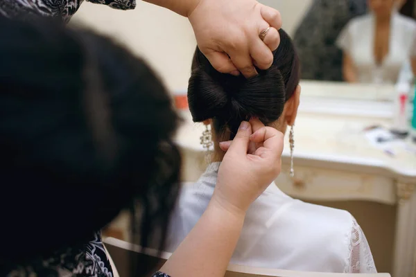 プロの美容師は 髪の弓を編むことによって美しいスレンダーなブルネットの花嫁の髪を作ります 白いコートを着た若い女性 朝の花嫁集まり — ストック写真