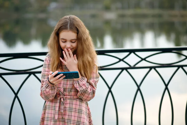 A girl with red hair in a pink checkered dress stands on the embankment and watches a funny video on her phone. Modern technologies in the mobile phone. Emotions of a child from watching video content
