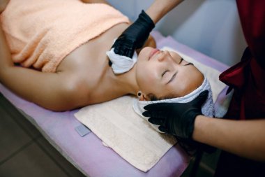 Kaplıcada kozmetik cilt bakımı. Modern kozmetoloji, eldivenli profesyonel bir kozmetik uzmanı genç bir kadının yüzüyle ameliyatlar yapar, kremi peçetelerle siler.