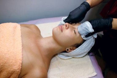 Kaplıcada kozmetik cilt bakımı. Modern kozmetoloji, siyah eldivenli profesyonel bir kozmetik uzmanı genç bir kızın yüzüyle ameliyatlar yapar, kremi peçetelerle siler.