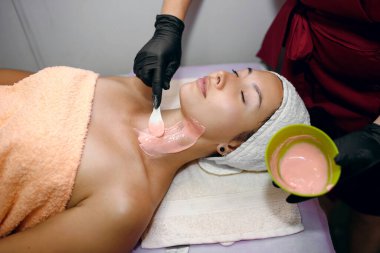 Güzel bir kız spa salonunda prosedürlere tabi tutuluyor, siyah eldivenli bir güzellik uzmanı pembe bir maske takıyor kalın bir tabakayla, derisini pislikten temizliyor, yüzünün saflığını gözlüyor.