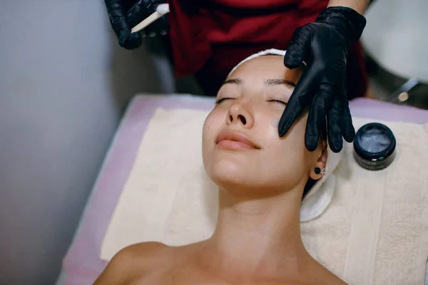 美しいブルネットはスパで手順を受けています マスター美容師は彼女の顔に保湿剤を適用します 顔のスキンケアの概念 プロのリフティング テキストのためのフリースペース — ストック写真