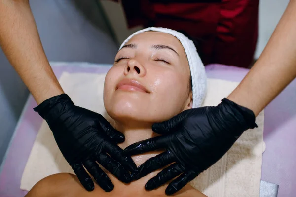 Güzellik merkezinde yüz masajı yaptıran genç bir kadının yakın çekimi. Cilt ve vücut bakımı için modern kozmetoloji. Siyah eldivenli profesyonel masör yüz masajı yapar.