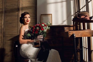 Kocaman bir buket kırmızı gül ve beyaz gelinlik içinde güzel bir genç kadın gelin antika mobilyalarla dolu bir odada oturuyor. Antika şapkalı eski tarz. Metin için boş alan