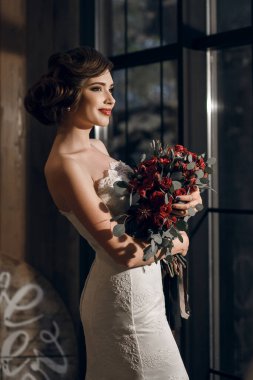 Beyaz gelinlik içinde güzel bir kadın gelin ve kocaman bir buket gülle beyaz eldivenler pencerenin yanında duruyor. Şık saç stili ve parlak kırmızı ruj. Kız mutlu ve gülümsüyor.