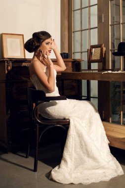 Beyaz bir gelinlik içinde, kozmetik ürünleri ve şık bir saç stili olan güzel bir gelin, antika mobilyalarla dolu bir odada oturuyor. Şapkalı eski tarz. Esmer, fotoğrafçı için poz veriyor.