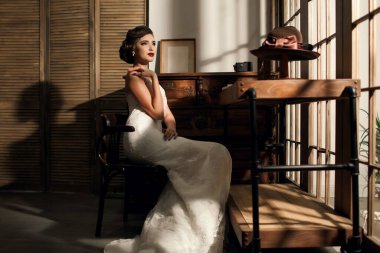 Makyajlı ve şık saç stili olan beyaz gelinlikli güzel bir gelin, antika mobilyalarla dolu bir odada oturuyor. Şapkalı eski tarz. Metin için yapay ışık ve boş alan