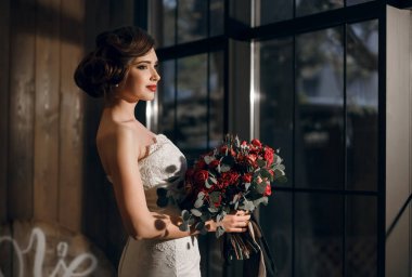 Pencerenin arka planında, beyaz gelinlik içinde güzel bir gelin-kız ve kırmızı gül buketli eldivenler var. Şık saç stili ve güzel makyaj. Metin için boş alan