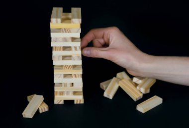 Siyah arka planda tahta masa oyunu. İnsanlar, dengenin önemli olduğu bir şans oyunu oynar. Tahta bloklardan yapılmış bir kule ve her seferinde bir parça koparan bir insan eli.