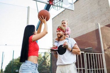 Genç sportif aile oyun sahasında basketbol oynuyor. Baba küçük kızını omuzlarına aldı, ve anne topu ringe fırlattı. Tarz sahibi aile gülüyor ve spor yapıyor.