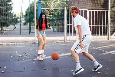Güzel, genç, atletik bir esmer erkek arkadaşıyla basketbol oynuyor ve topu almaya çalışıyor. Gençler eğleniyor ve spor yapıyor. Kot şortlu ve kırmızı tişörtlü bir kız.