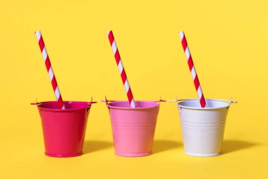 Sarı arka planda üç küçük kova ve çizgili pipetler. Kovalar kırmızı ve beyaz. Uçsuz bucaksız susuzluk ve sıcak bir günde suya ihtiyaç duyma kavramı. Sosyal ağlar için parlak fotoğraf