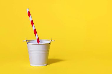 Sarı arka planda beyaz bir oyuncak kovası ve şeritli kırmızı ve beyaz bir pipet. İlginç ve alışılmadık bir sunumda lezzetli bir içecek konsepti. Metin için boş alan