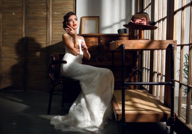 Beyaz bir gelinlik içinde güzel bir esmer gelin şapkalı ve antika mobilyalı klasik bir iç mekanda oturuyor. Düğün gününde, genç ve güzel bir kadın düğün fotoğrafçısı için poz verir.
