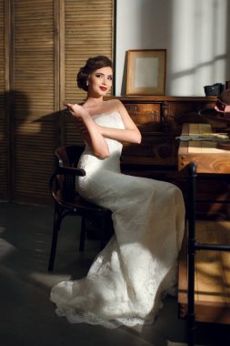 Beyaz bir gelinlik içinde güzel bir esmer gelin şapkalı ve antika mobilyalı klasik bir iç mekanda oturuyor. Genç ve güzel bir kadın bir düğün fotoğrafçısı için poz veriyor. Gelinin dikey fotoğrafı.