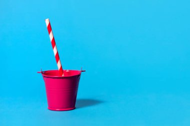 Açık mavi arka planda kırmızı ve pembe bir oyuncak kovası ve şeritli kırmızı ve beyaz bir pipet. Standart olmayan içki servisi yapan bir parti ya da sıcak bir günde suya ihtiyaç duyan bir parti.