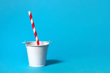 Açık mavi arka planda beyaz bir oyuncak kovası çizgili kırmızı ve beyaz bir pipet içiyor. Hafta sonu parti konsepti. Standart olmayan içecekler ve susuzluk. Metin için boş alan
