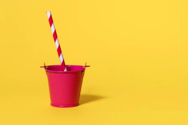 Sarı arka planda kırmızı-beyaz şeritli pipetli kırmızı oyuncak kovası. Uçsuz bucaksız susuzluk ve sıcak bir günde suya ihtiyaç duyma kavramı. Metin için boş alan