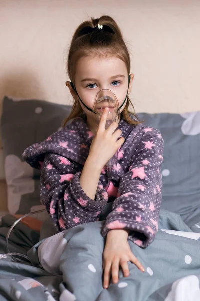 A little girl sits on the bed and is treated at home with an inhaler. Allergies and respiratory diseases. Treatment of a child with an inhaler at home under the supervision of parents vertical photo